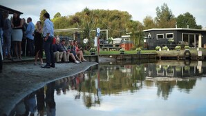 A group of people by a calm water body, a dock, and a building with a flat roof.