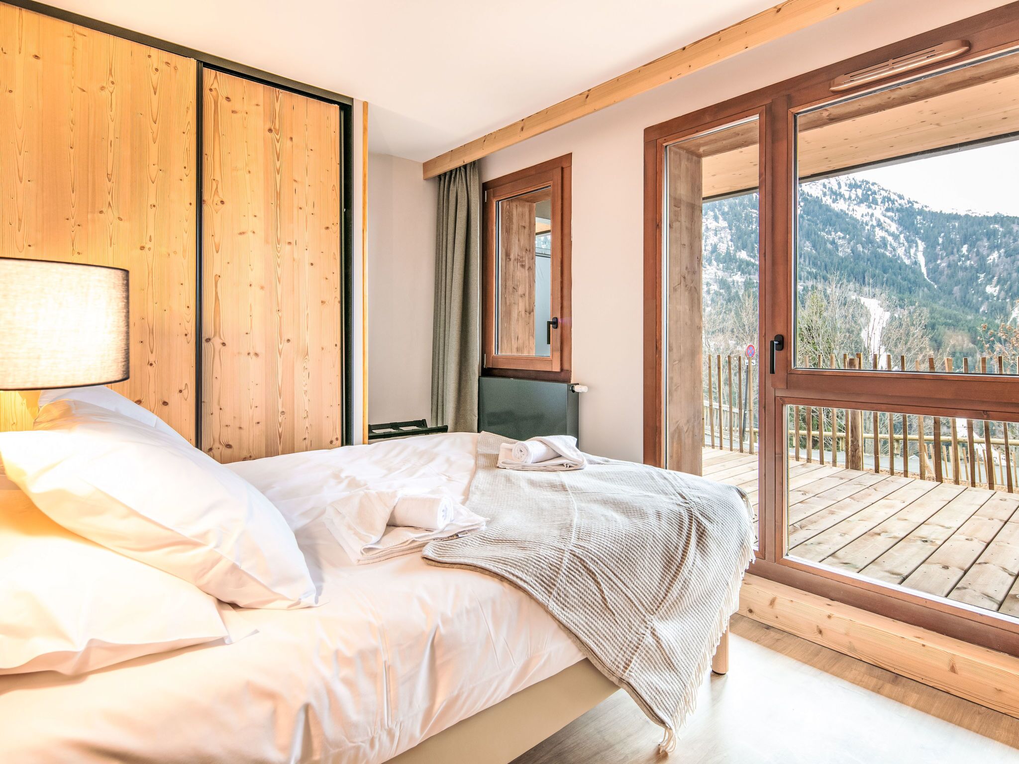 A bedroom with a bed, wooden walls, a window, and a balcony with a view of mountains.