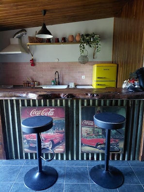 A kitchen with a yellow appliance, two stools with Coca-Cola posters, a hanging plant, and a brick wall backsplash. - La Halte de Dakota, a unique bed-and-breakfast nestled in the heart of nature. (Chamouillac)