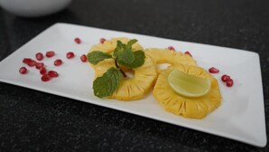 A white rectangular plate with pineapple slices, lime wedges, mint leaves, and pomegranate seeds on a dark countertop.