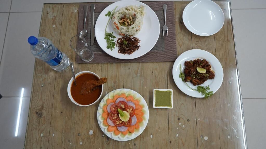 A wooden table with a meal consisting of rice, a meat dish, a vegetable dish, a bowl of sauce, and a small dish of green chutney.