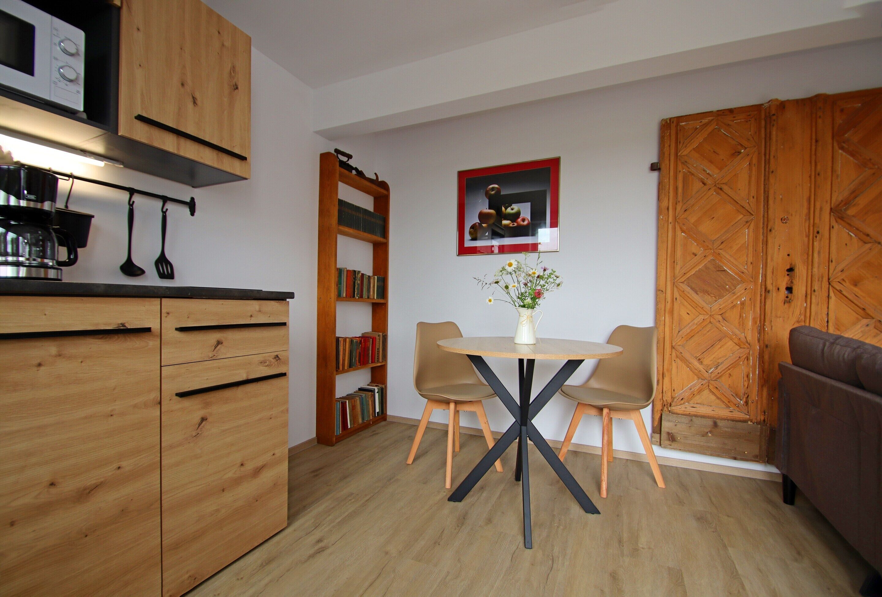 A compact kitchen with wooden cabinets, a dining table with chairs, a bookshelf, and a wooden wardrobe.