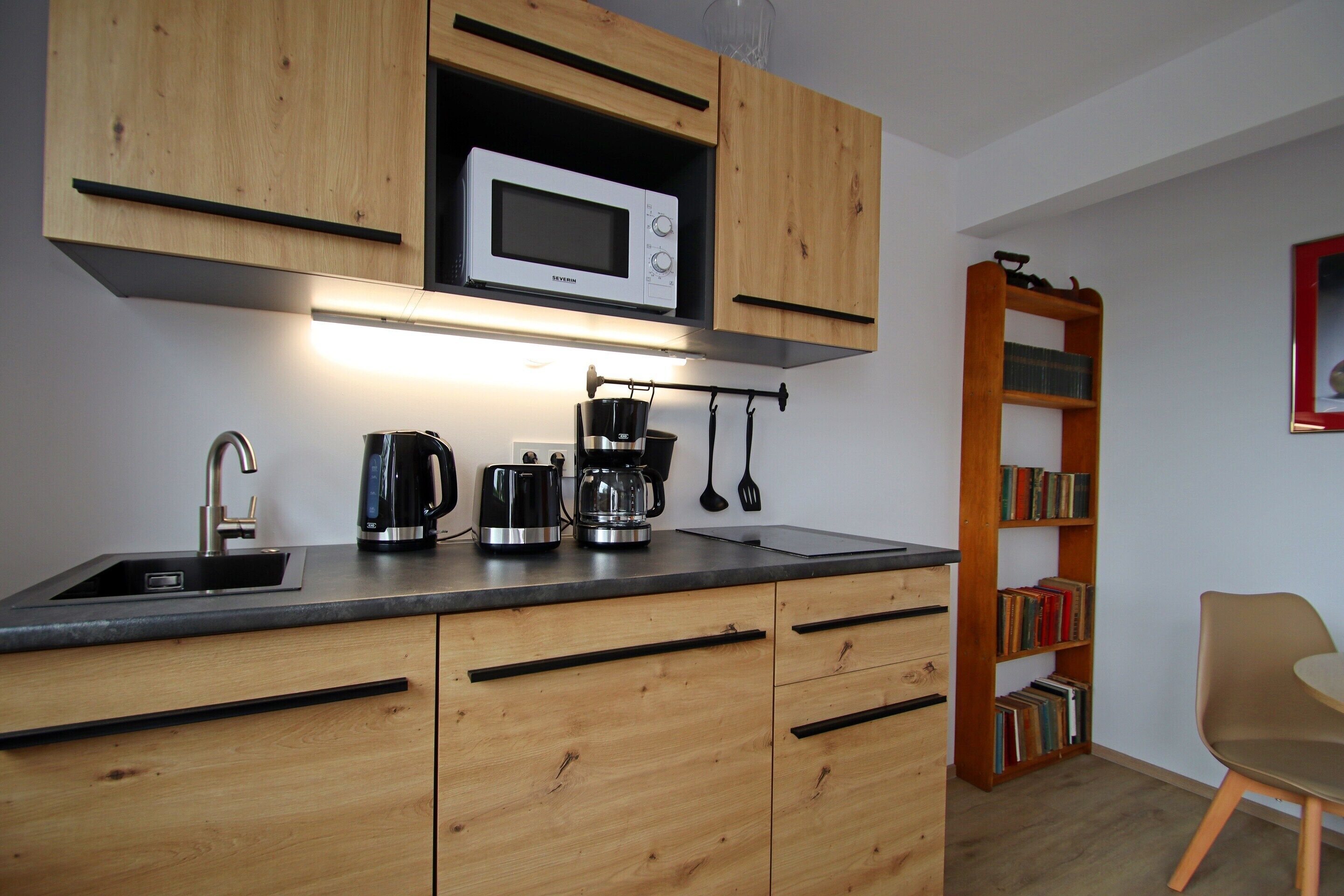 A kitchen with wooden cabinets, a microwave, a coffee maker, and a bookshelf.