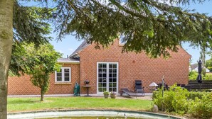 Una casa in mattoni con un'ampia finestra, un piccolo patio con tavolo e sedie, e uno stagno ricoperto di ninfee.
