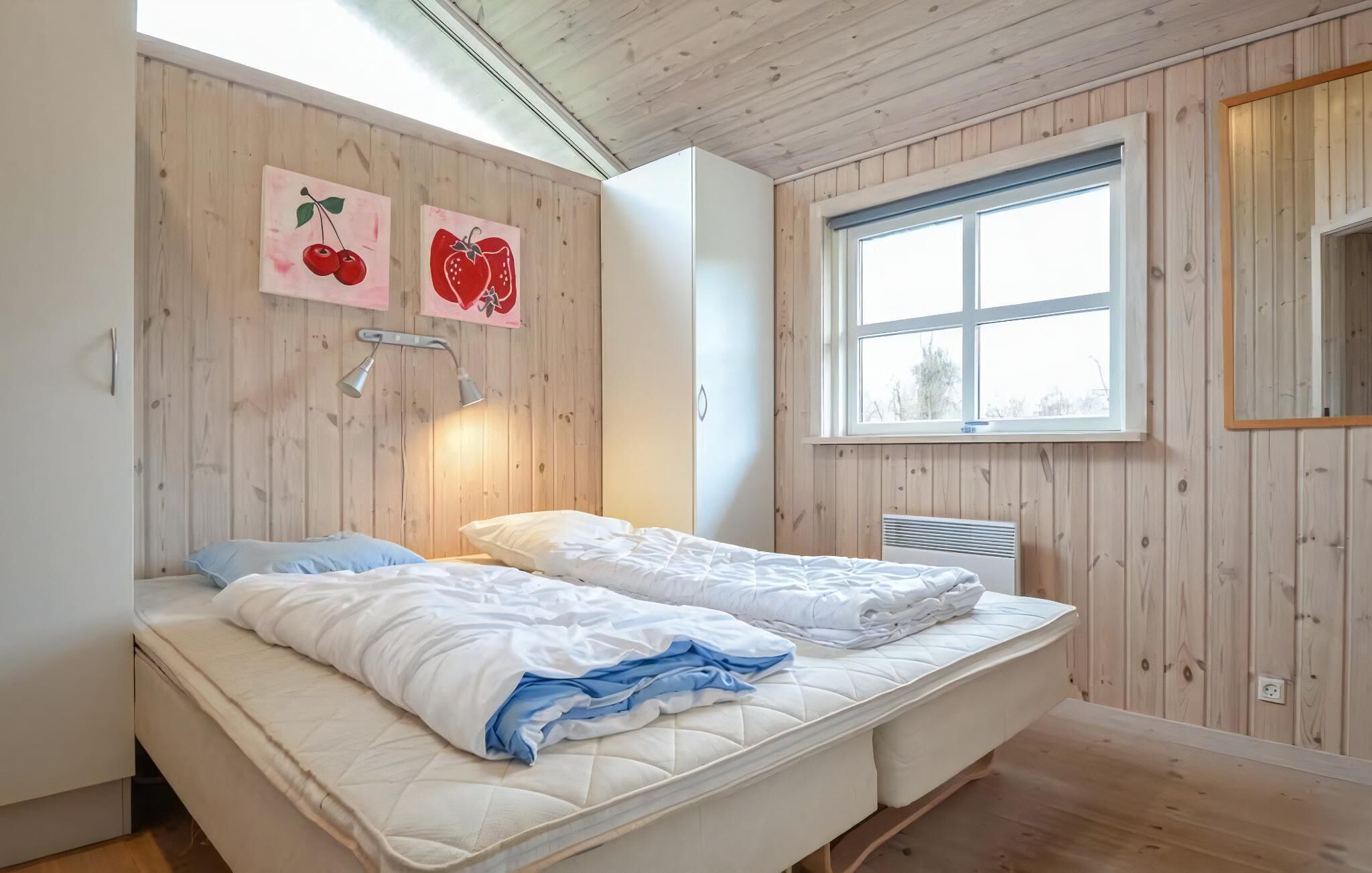 Ein Zimmer mit Holzwänden, einem Bett mit weißer Bettwäsche, einem Fenster und zwei an der Wand hängenden Stillleben mit Früchten.