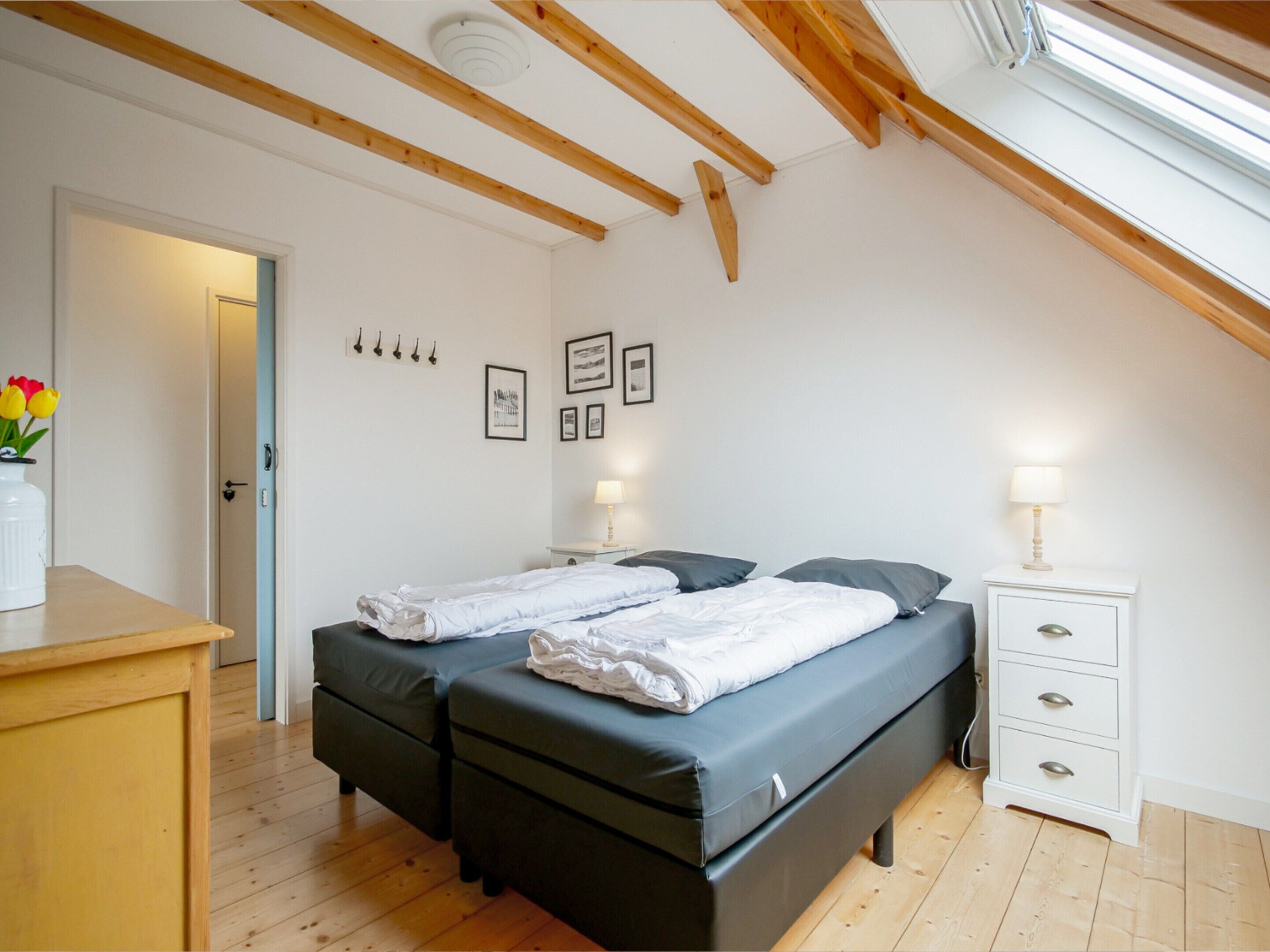 Un dormitorio con dos camas, techo de madera, pared blanca con cuadros enmarcados, mesita de noche con lámpara y cómoda de madera.