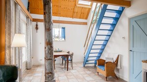 A loft-style room with a sloped ceiling, exposed wooden beams, a dining area, a blue staircase, and a black leather sofa. - Cozy Vacation Rental in a Former Farmhouse, Surrounded by Nature (Callantsoog)