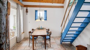 A dining area with a wooden table and chairs, a window with blue curtains, a hanging light fixture, and a blue staircase. - Cozy Vacation Rental in a Former Farmhouse, Surrounded by Nature (Callantsoog)