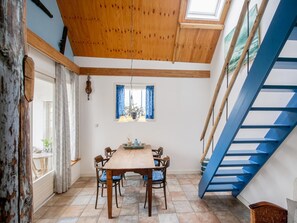 Uma área de refeições com mesa e cadeiras de madeira, janela com cortinas azuis, luminária pendente e escada azul.