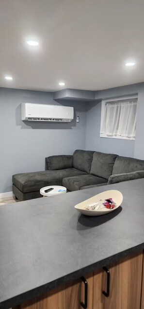 A modern living room with a grey sofa, a small round table, and an air conditioning unit mounted on the wall. - Salt and Spruce private 1 bedroom suite with double bed, kitchen, and laundry (Mount Pearl)