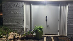A grey door with a keypad lock, flanked by two potted plants with purple flowers, set against a textured wall. - Salt and Spruce private 1 bedroom suite with double bed, kitchen, and laundry (Mount Pearl)