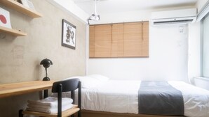 Een slaapkamer met een bed, een bureau, een stoel, een plank met een afbeelding en een raam met jaloezieën.