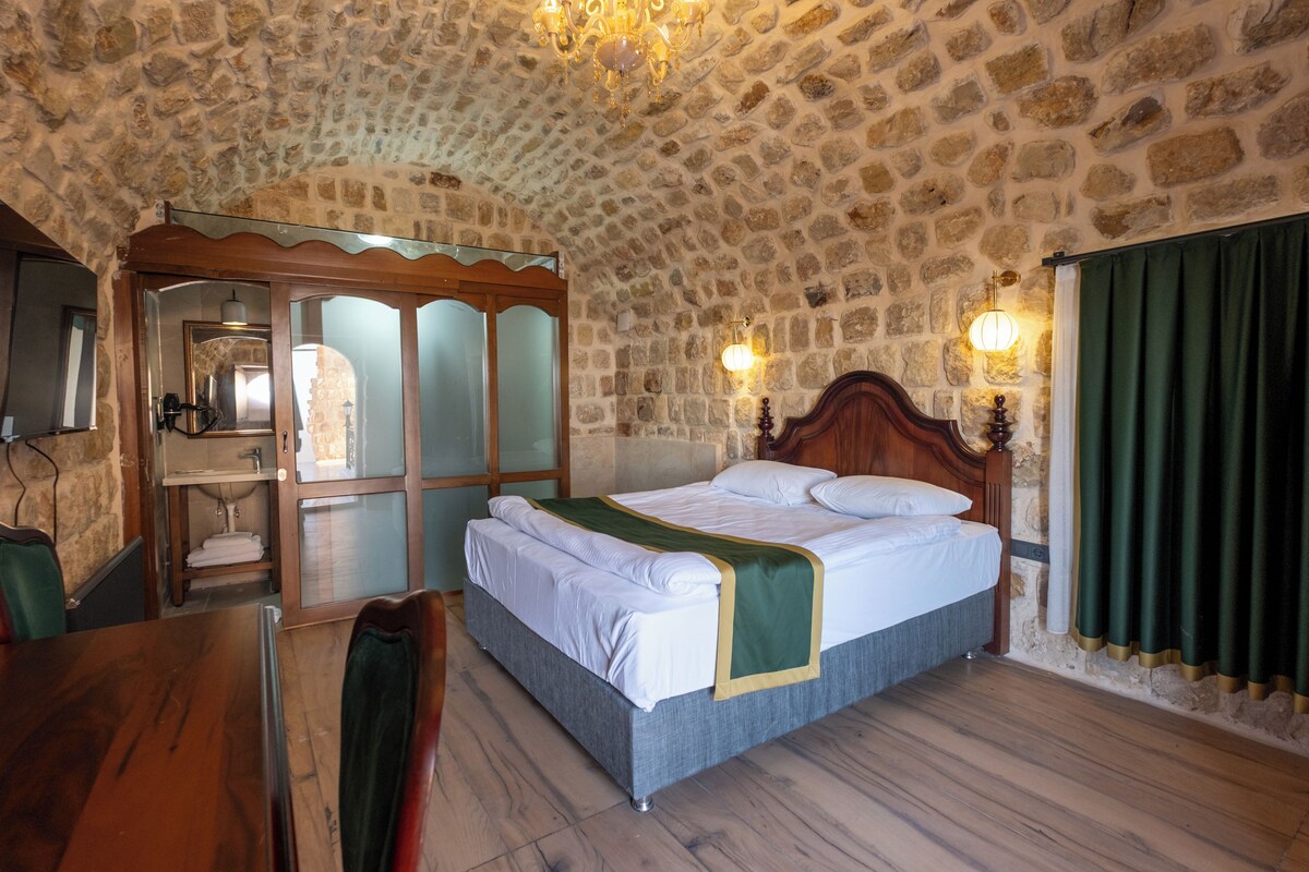 A stone-walled room with a bed, a wooden chair, a table, and a mirror.