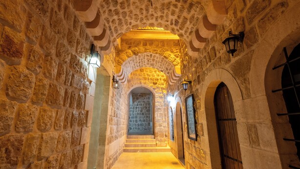 Un pasillo de piedra con arcos, una puerta de madera y faroles en la pared.