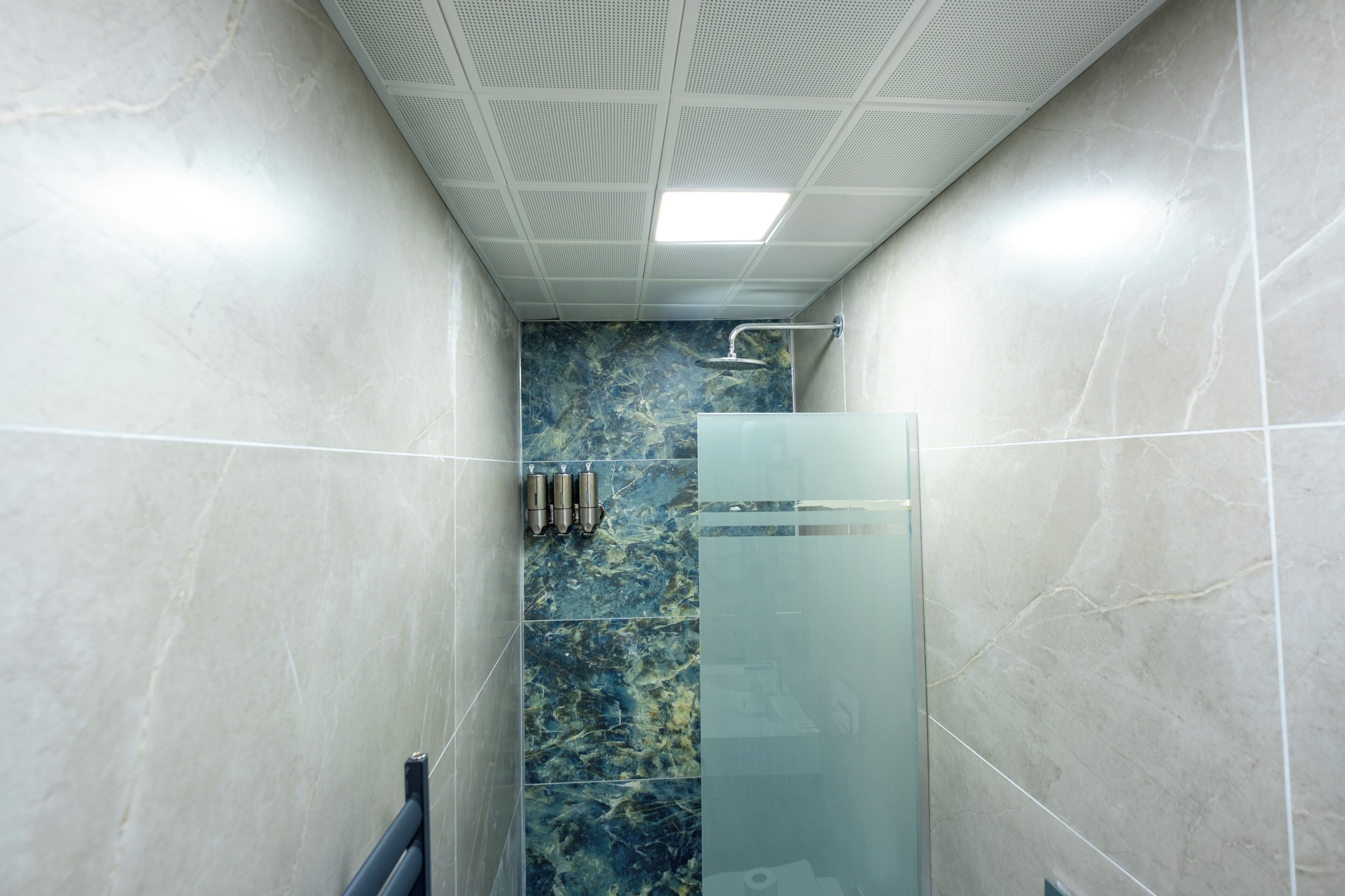 A narrow bathroom with a glass shower enclosure, marble tiles, and a ceiling with a grid pattern.