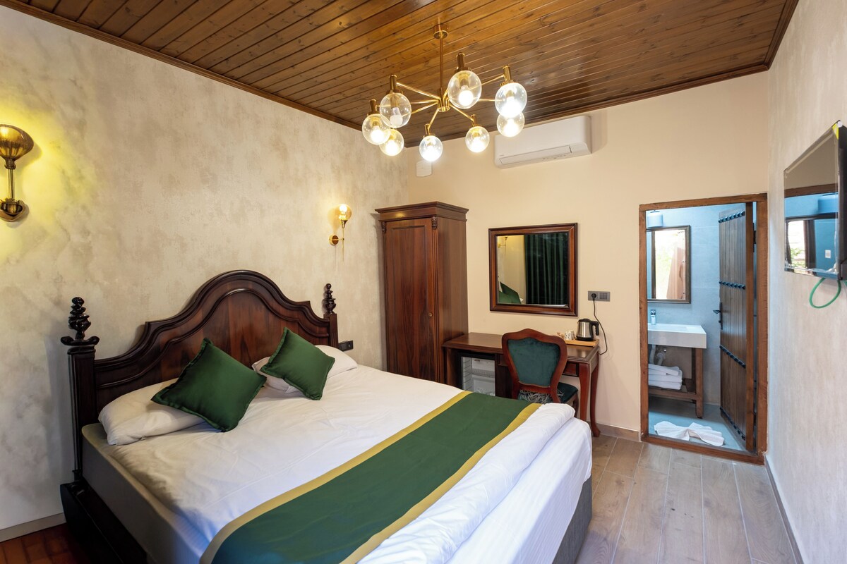 A bedroom with a wooden ceiling, a bed with green and white bedding, a desk with a chair, a mirror, and a bathroom visible through an open door.