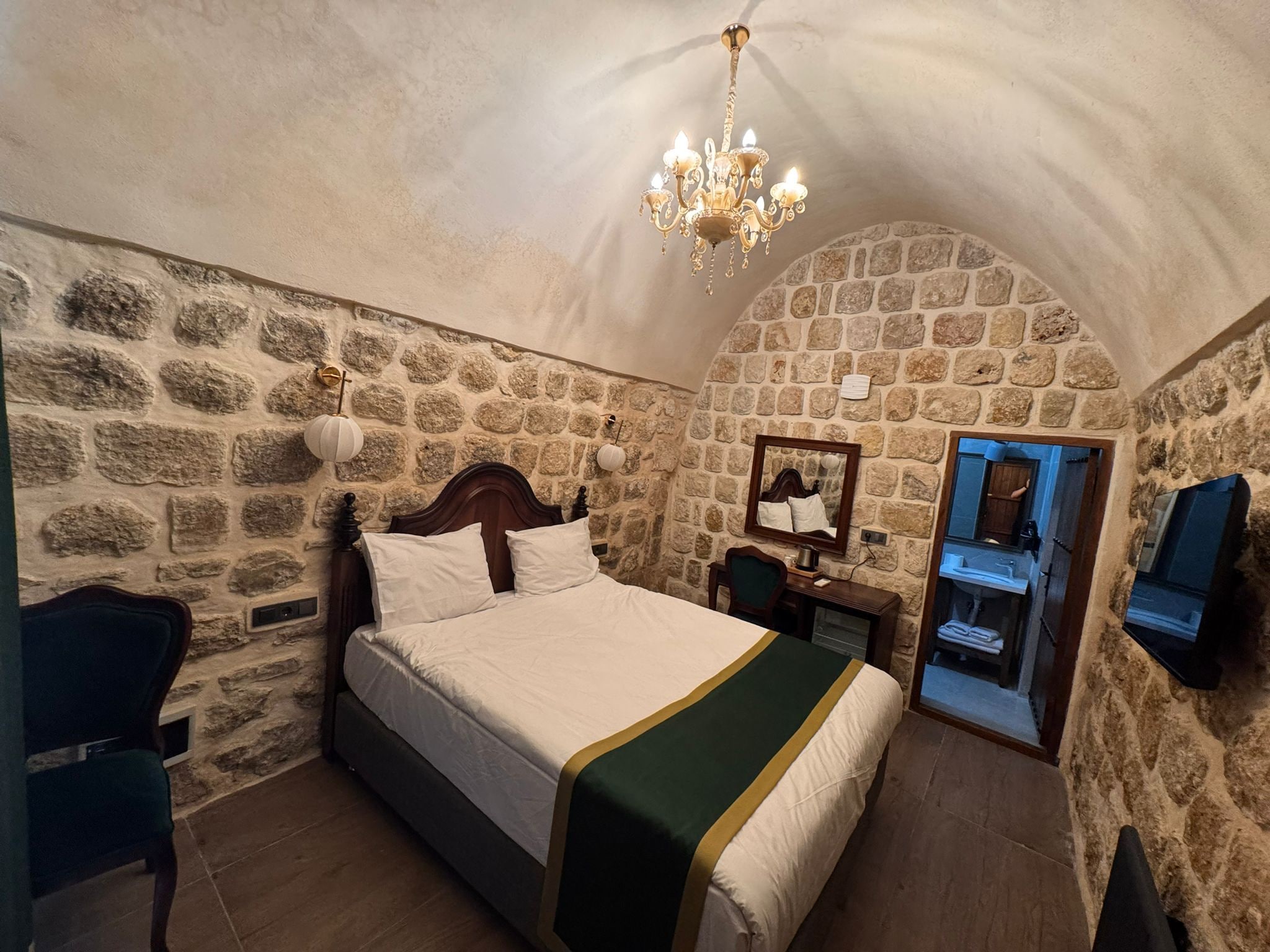 A bedroom with a stone wall, a bed with white bedding, a wooden nightstand, a chair, a mirror, and a chandelier.