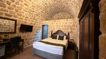 A bedroom with a stone wall, a bed with green and white bedding, a desk with a chair, a television, and a wooden wardrobe.