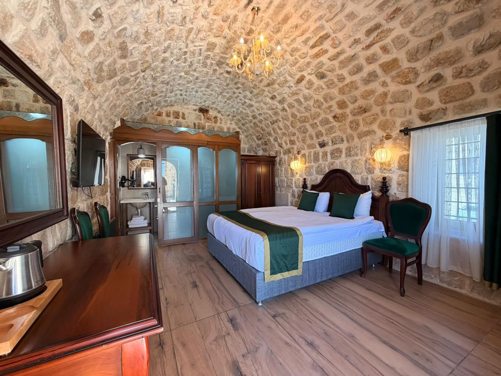 A room with a stone ceiling, a bed with green and white bedding, a wooden desk, and a chair.