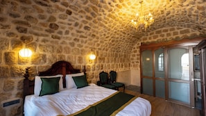 Quarto com paredes de pedra, uma cama, duas cadeiras, um candelabro e uma porta com painéis de vidro.