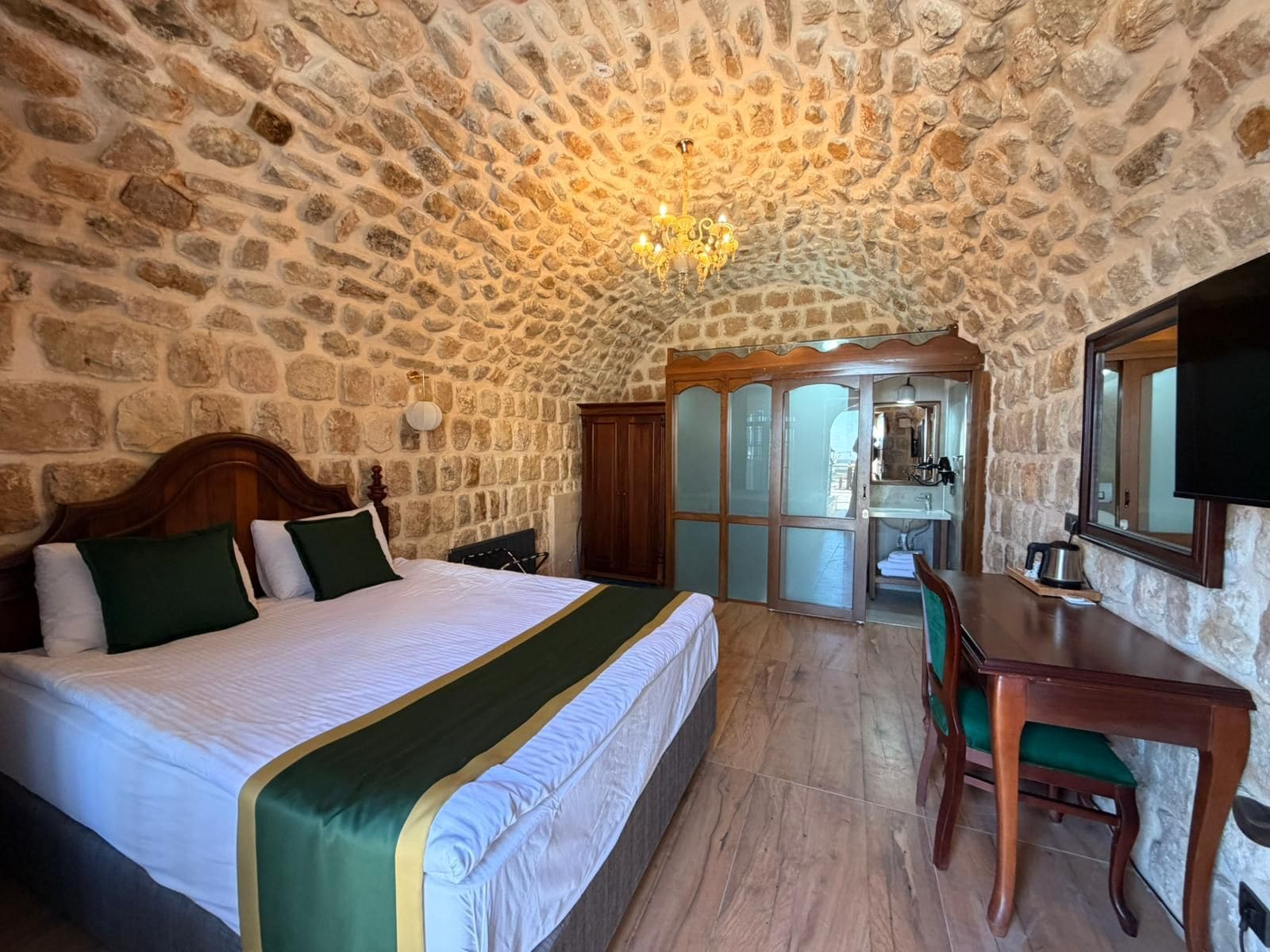 A bedroom with a stone wall, a bed with green and white bedding, a wooden desk, and a chandelier.