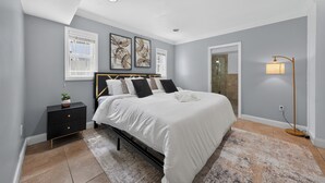 Een slaapkamer met een bed, nachtkastje, lamp, kunstwerk en een raam.