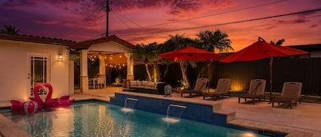 Ein Schwimmbecken mit Liegestühlen und Sonnenschirmen, ein kleines Haus mit einer Tür sowie ein Sonnenuntergangshimmel.