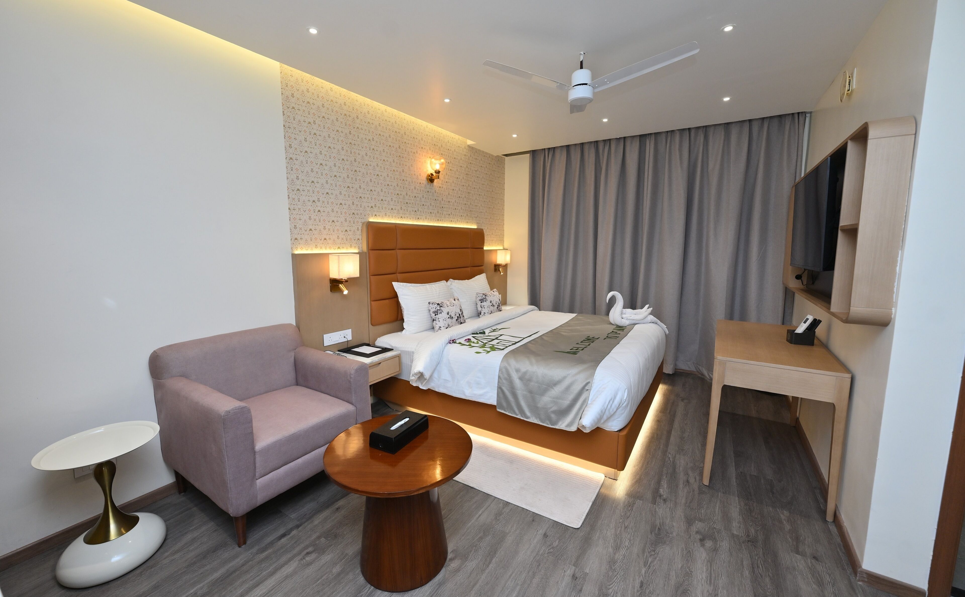 A modern hotel room with a bed, a desk, a chair, a small table, a TV, and a ceiling fan.
