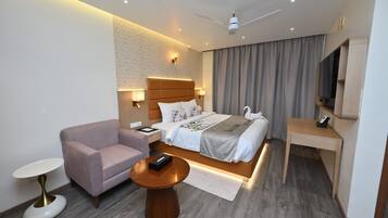 A modern hotel room with a bed, a desk, a chair, a small table, a TV, and a ceiling fan.