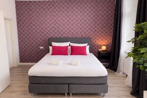 A neatly made bed with white and red pillows, a grey upholstered headboard, and a bedside table with a lamp. - MIILOX Apartments Dresden am Park (Dresden)