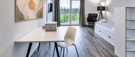Habitación de hotel moderna con escritorio blanco, laptop, silla, cama, cómoda y ventana grande con vista a follaje.