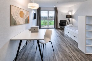 Habitación de hotel moderna con escritorio blanco, laptop, silla, cama, cómoda y ventana grande con vista a follaje.