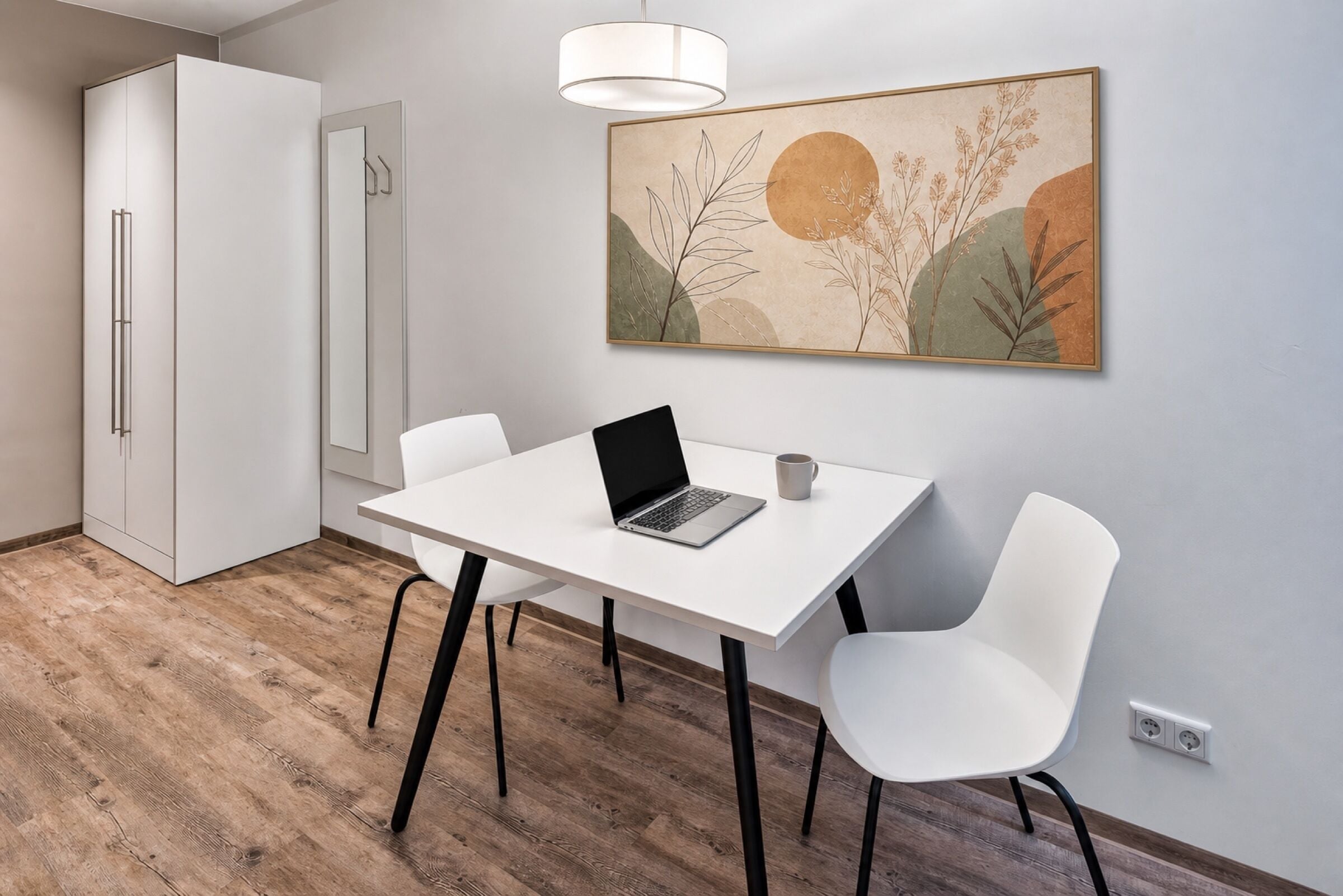 Une cuisine moderne avec des meubles blancs, une table à manger avec deux chaises, un ordinateur portable posé sur la table et un luminaire fixé au mur.