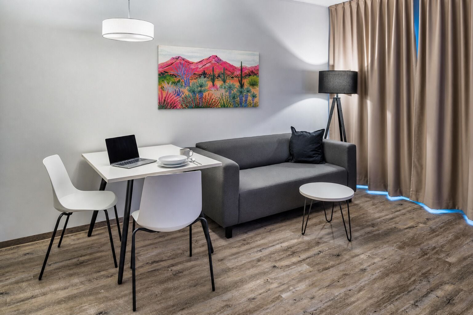 Une pièce moderne avec un canapé gris, un bureau blanc avec un ordinateur portable, deux chaises blanches, une petite table ronde, un lampadaire et un tableau représentant un paysage désertique.