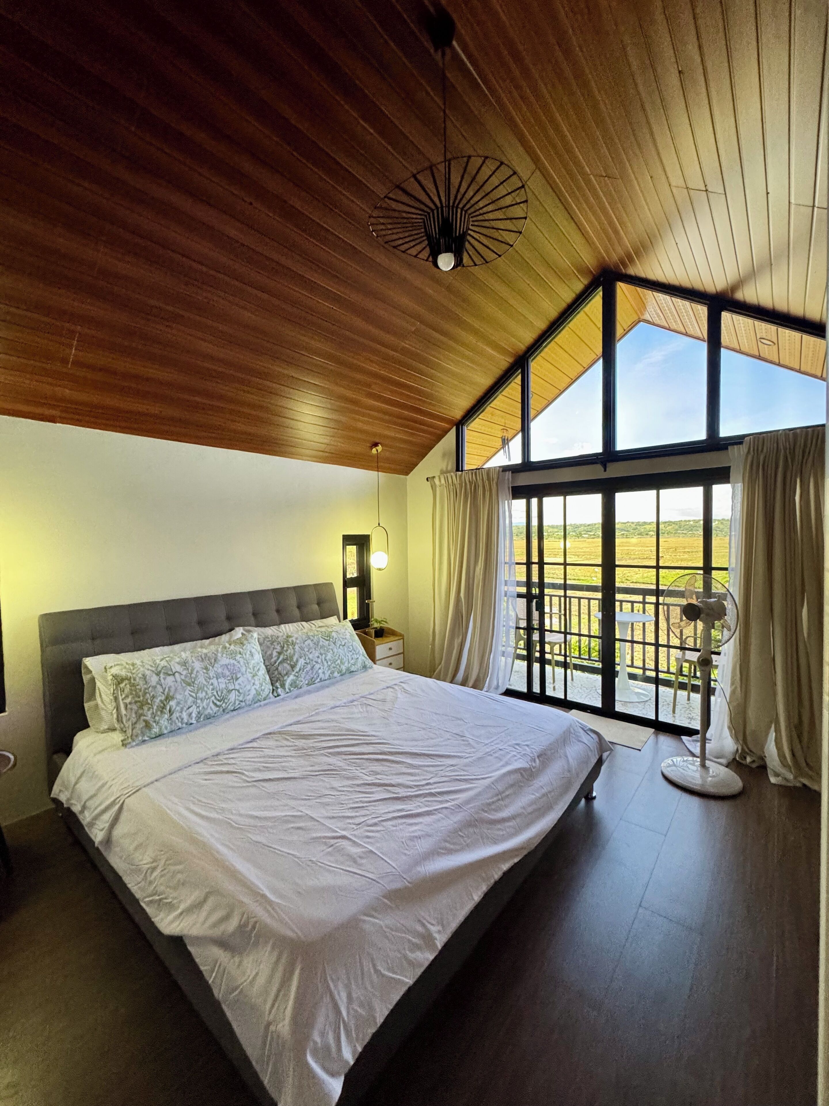 Une chambre dotée d’un grand lit, d’un plafond en bois et offrant une vue sur un paysage verdoyant grâce à de grandes fenêtres.