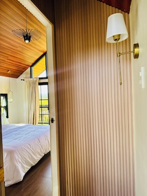 A hotel room with a wooden ceiling, a bed with white bedding, a window with curtains, and a wall-mounted lamp.