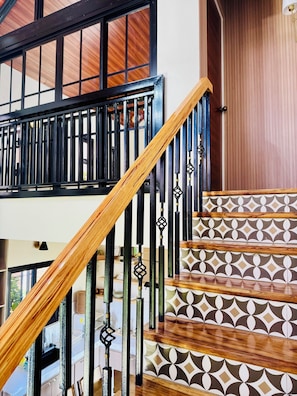 A staircase with a wooden handrail and black metal balusters, leading to a room with a patterned floor and a wooden door.