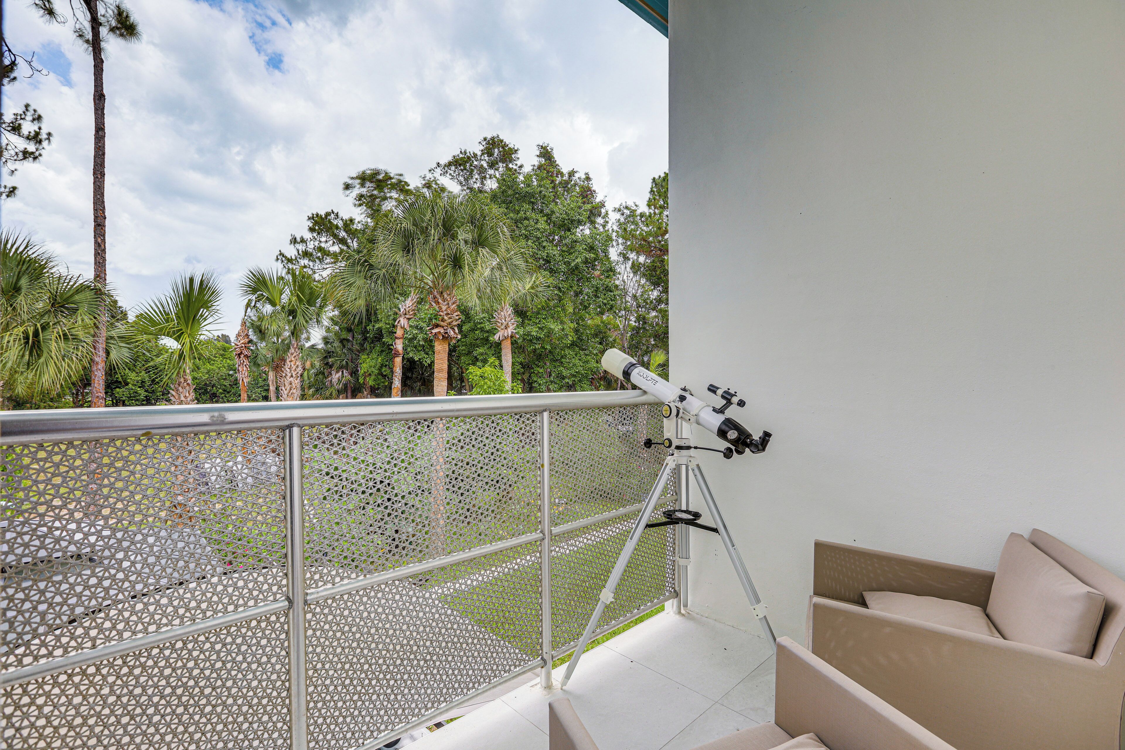 A balcony with a telescope, a white chair, and a view of palm trees and a clear sky.