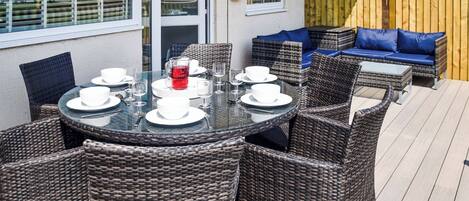 Eine Terrasse mit Korbmöbeln, einem Glastisch gedeckt für ein Essen und einem Holzzaun.