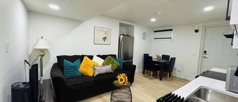 En moderne stue med en sort sofa, et sofabord med en vase solsikker, et spiseområde med et sort bord og stole, og et køkken med vask og komfur.