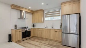 A modern kitchen with wooden cabinets, a stainless steel refrigerator, a stove, and a window with blinds. - Pavilion Lakefront Luxe 3 Bdrm near Lake! (Chestermere)