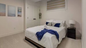 A bedroom with a bed, a nightstand, a lamp, a door, and wall art. - Pavilion Lakefront Luxe 3 Bdrm near Lake! (Chestermere)