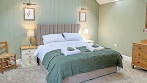 Ein Schlafzimmer mit schrägen Wänden, einem Bett mit grüner Bettdecke, zwei Nachttischlampen, einem Holzstuhl und einer Kommode.