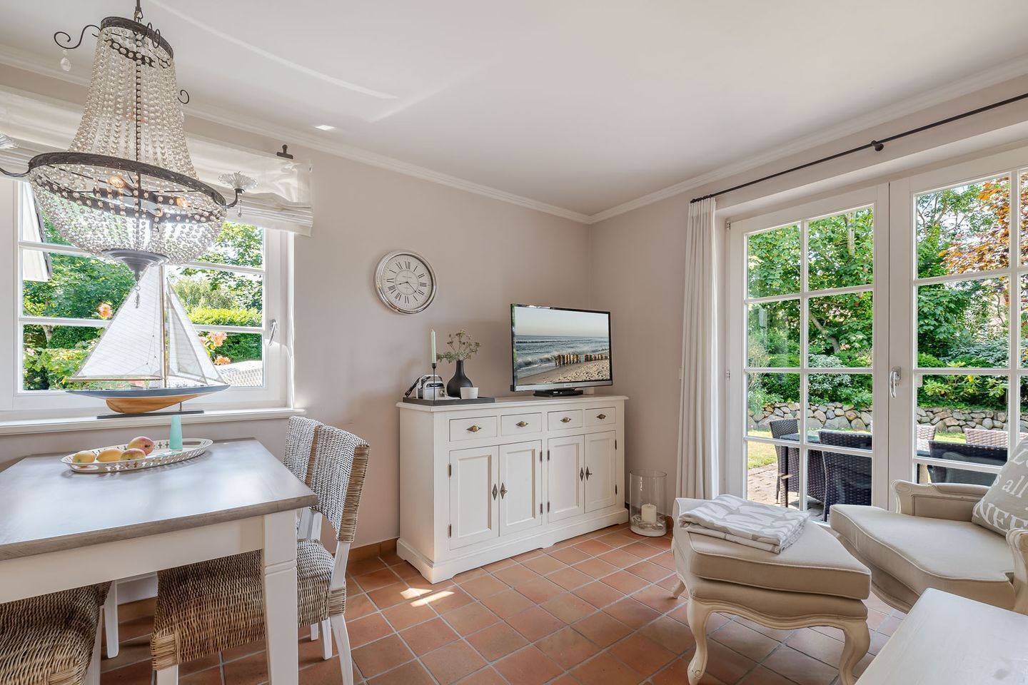 A room with a dining table, chairs, a television, a cabinet, and large windows with a view of greenery.