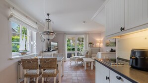 Una cocina con armarios blancos, un comedor con sillas de mimbre, una araña y vistas amplias a través de grandes ventanas.