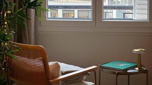 A room with a window overlooking a cityscape, a chair, a small table with a lamp, and a plant. - Charming Parisian Retreat with Eiffel Tower Views (Paris)