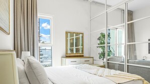 A bedroom with a large bed, a nightstand, a dresser, a mirror, and a window with a view of the city. - Bloomfields 1BR - Golf Ville - DXB Hills (Dubai)