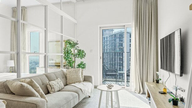 A modern living room with a sofa, a small round table, a TV, and a balcony with a view of buildings. - Bloomfields 1BR - Golf Ville - DXB Hills (Dubai)