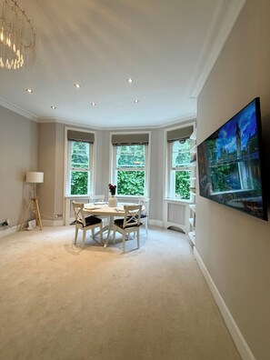 A dining room with a table, chairs, and a chandelier. There are three windows with views of greenery. - Luxury Apartment Overlooking St George’s Square & the Thames (London)