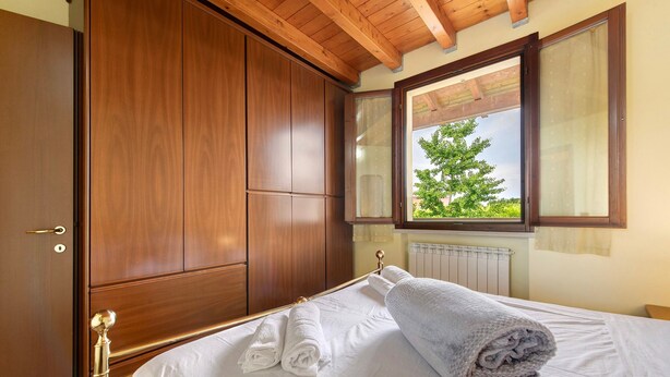 Une chambre à coucher avec une armoire en bois, un lit recouvert de draps blancs et une fenêtre donnant sur des arbres.